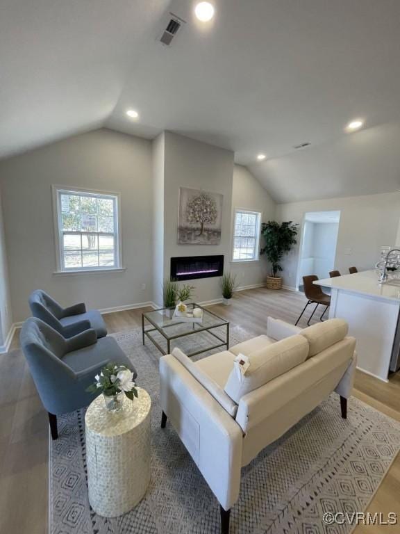 living room featuring vaulted ceiling, a warm lit fireplace, wood finished floors, and visible vents