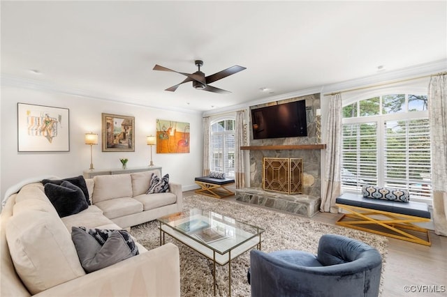 living area with ceiling fan, a stone fireplace, wood finished floors, baseboards, and crown molding