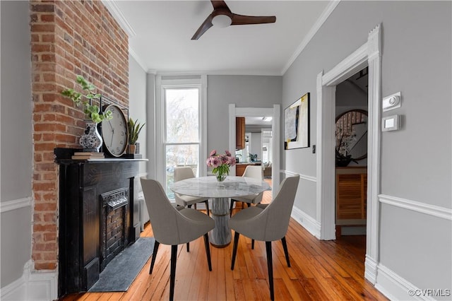 dining room featuring hardwood / wood-style floors, baseboards, ceiling fan, ornamental molding, and a brick fireplace