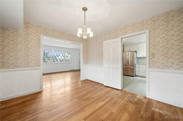 empty room with a wainscoted wall, light wood-style flooring, wallpapered walls, and an inviting chandelier