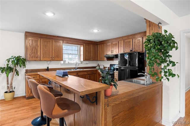 kitchen featuring a breakfast bar area, a sink, black appliances, light wood-style floors, and under cabinet range hood