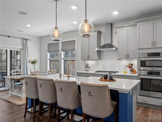 kitchen featuring wall chimney range hood, dark wood finished floors, a center island with sink, light countertops, and appliances with stainless steel finishes