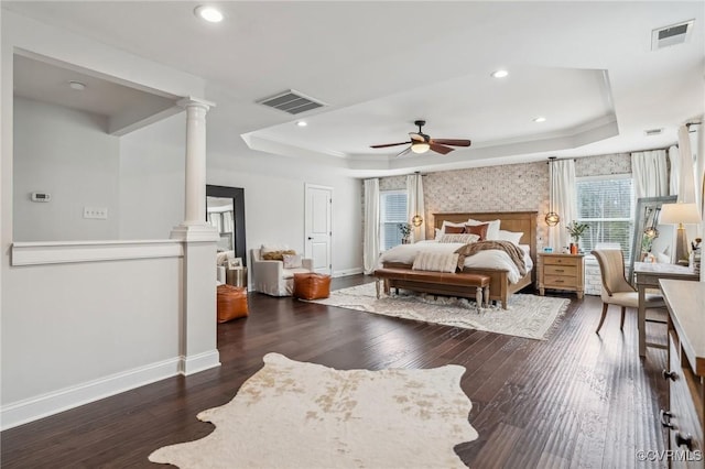 bedroom with a tray ceiling, wood finished floors, visible vents, and ornate columns