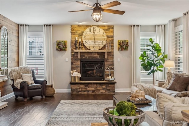 living area featuring a stone fireplace, baseboards, a ceiling fan, and dark wood-style flooring