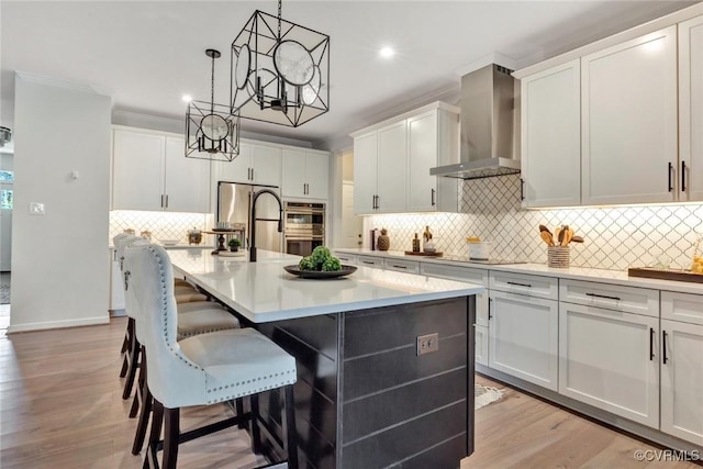 kitchen with a center island with sink, stainless steel appliances, hanging light fixtures, light countertops, and wall chimney range hood