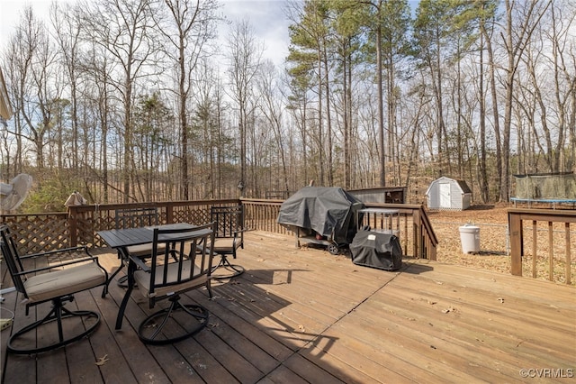 wooden terrace featuring outdoor dining space, an outbuilding, a shed, a trampoline, and a grill