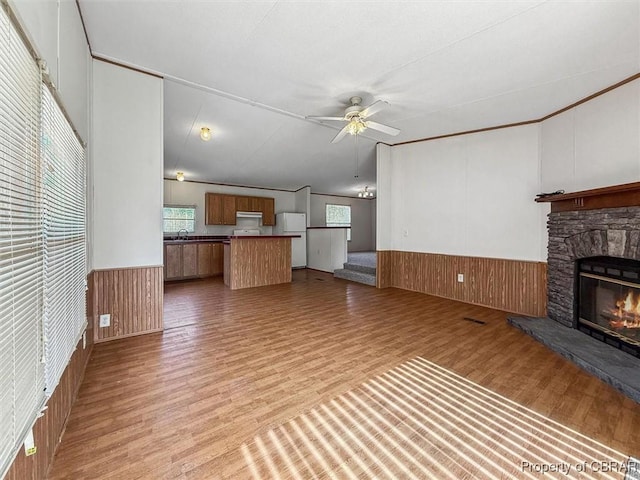 unfurnished living room featuring wooden walls, ceiling fan, a wainscoted wall, a stone fireplace, and light wood-style flooring