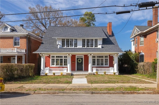 view of front of house featuring a chimney, covered porch, a front lawn, and a shingled roof