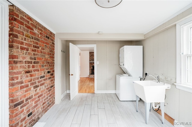 laundry room featuring light wood-style floors, brick wall, stacked washer / drying machine, baseboards, and laundry area