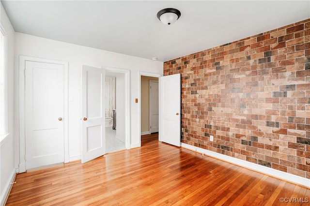 unfurnished bedroom with brick wall, baseboards, light wood finished floors, and connected bathroom