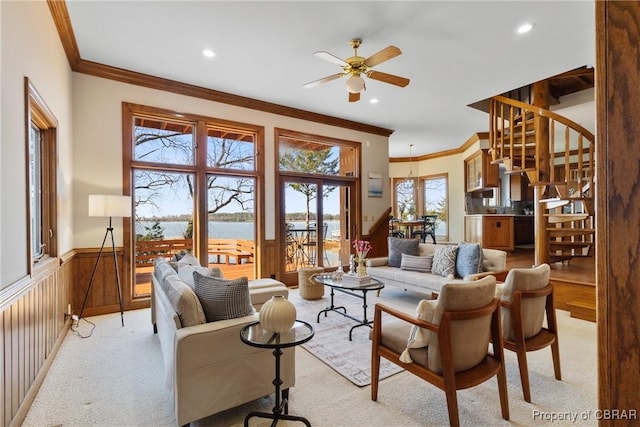 living room featuring a wainscoted wall, a water view, stairway, crown molding, and light colored carpet