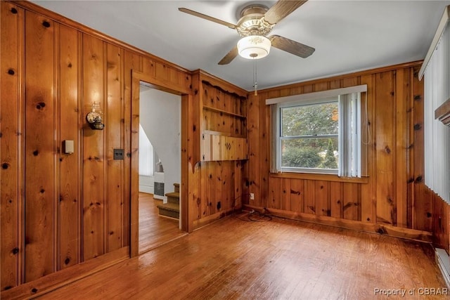 unfurnished room featuring light wood-type flooring, stairway, wooden walls, baseboard heating, and ceiling fan