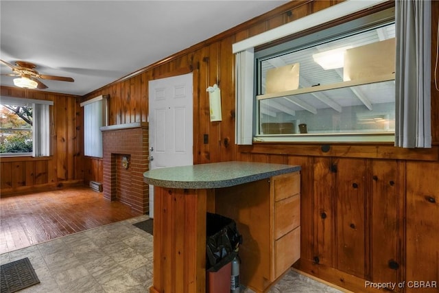 kitchen with a brick fireplace, wood walls, brown cabinetry, a ceiling fan, and a baseboard radiator