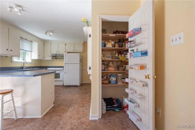 kitchen with dark countertops, under cabinet range hood, a kitchen breakfast bar, white appliances, and a sink