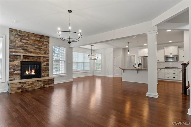 unfurnished living room featuring a stone fireplace, dark wood-type flooring, baseboards, and ornate columns