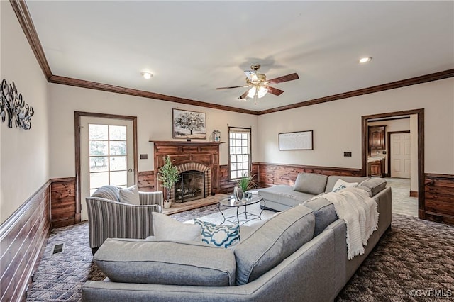 living room featuring ornamental molding, carpet floors, wainscoting, a brick fireplace, and ceiling fan