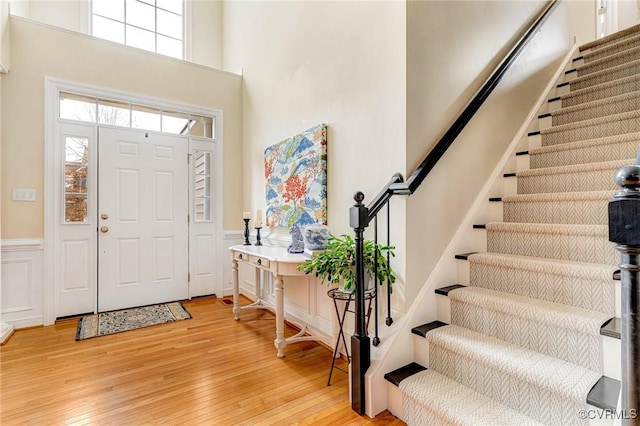 foyer featuring stairway, light wood-style floors, a towering ceiling, wainscoting, and a decorative wall