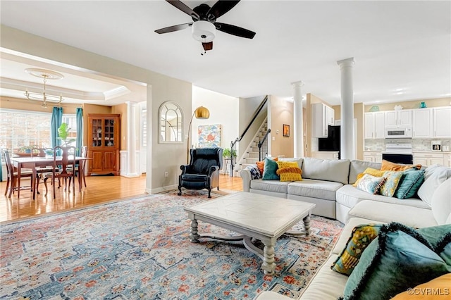 living area with ornate columns, a tray ceiling, light wood-style flooring, ceiling fan, and stairs