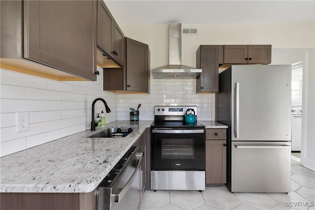 kitchen with dark brown cabinetry, light stone counters, appliances with stainless steel finishes, wall chimney exhaust hood, and a sink