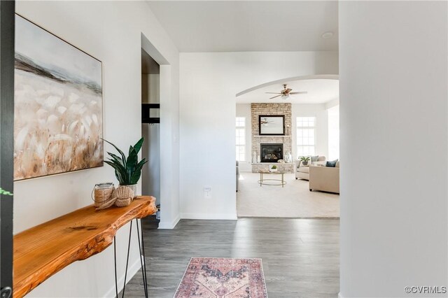 interior space with a ceiling fan, wood finished floors, arched walkways, a stone fireplace, and baseboards