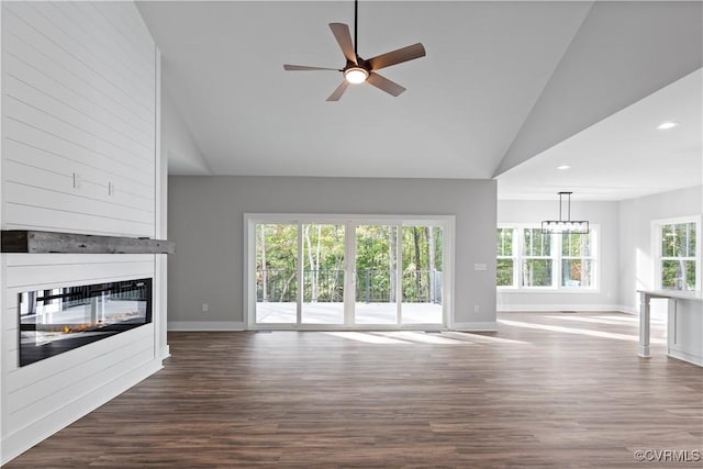 unfurnished living room with baseboards, high vaulted ceiling, dark wood-type flooring, a glass covered fireplace, and ceiling fan with notable chandelier