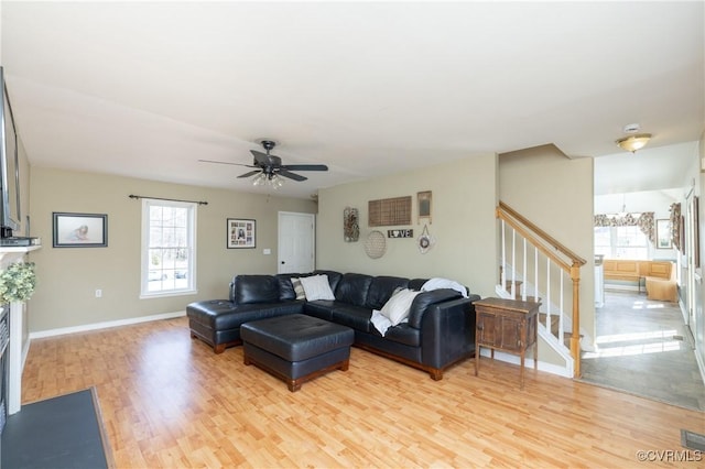 living area with stairway, baseboards, a fireplace, light wood-style floors, and ceiling fan with notable chandelier