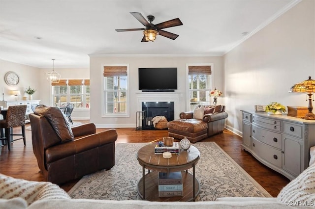 living area featuring a fireplace with flush hearth, ornamental molding, ceiling fan with notable chandelier, dark wood-style floors, and baseboards