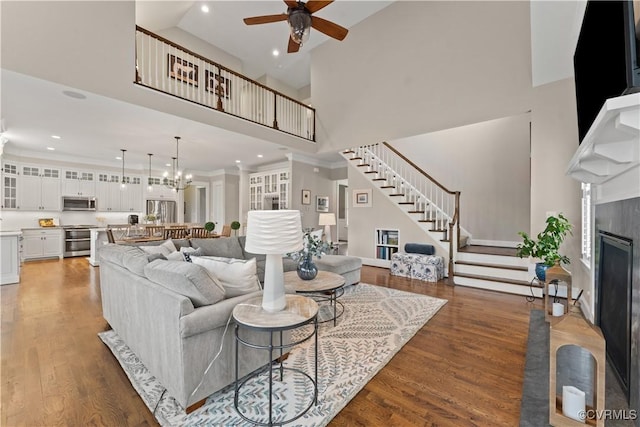 living area featuring wood finished floors, a high ceiling, a fireplace with flush hearth, stairs, and ceiling fan with notable chandelier