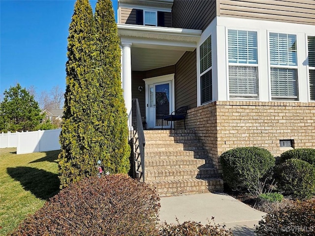 view of exterior entry with a lawn, fence, brick siding, and crawl space