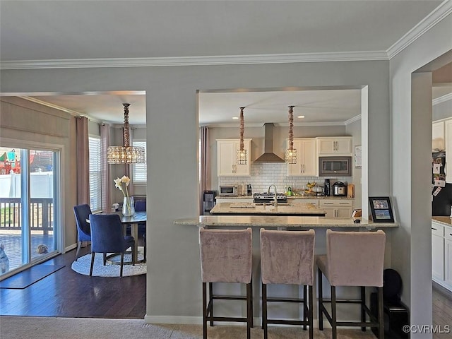 kitchen with ornamental molding, a kitchen bar, stainless steel microwave, wall chimney range hood, and backsplash