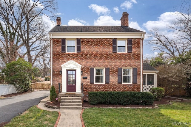 colonial house featuring a front yard, fence, brick siding, and a chimney