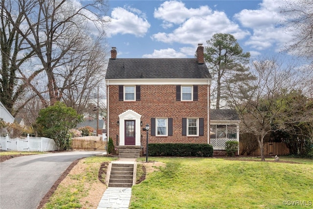 colonial inspired home with a front yard, fence, a chimney, aphalt driveway, and brick siding