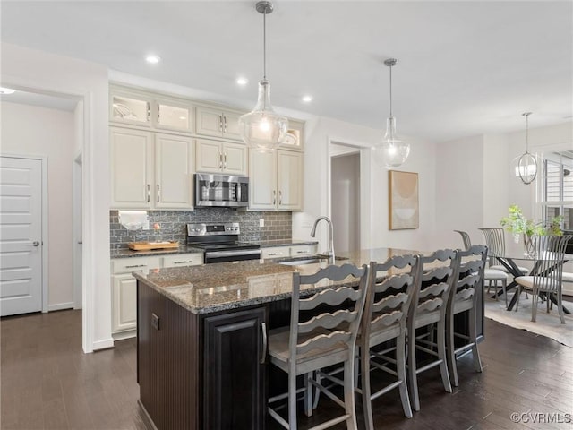 kitchen featuring a center island with sink, dark stone counters, a sink, stainless steel appliances, and backsplash