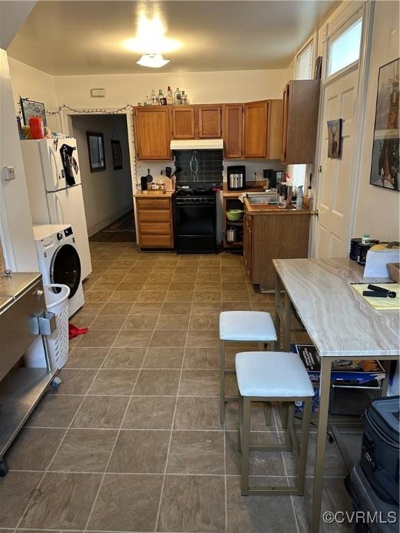 kitchen with brown cabinetry, washer / clothes dryer, tile patterned flooring, black gas range, and under cabinet range hood