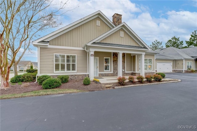 craftsman-style home with aphalt driveway, covered porch, a chimney, a garage, and stone siding