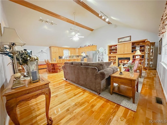 living area with light wood finished floors, a warm lit fireplace, lofted ceiling with beams, and ceiling fan