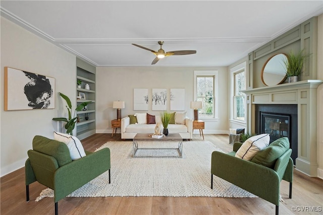 living room featuring built in shelves, a ceiling fan, wood finished floors, baseboards, and a glass covered fireplace