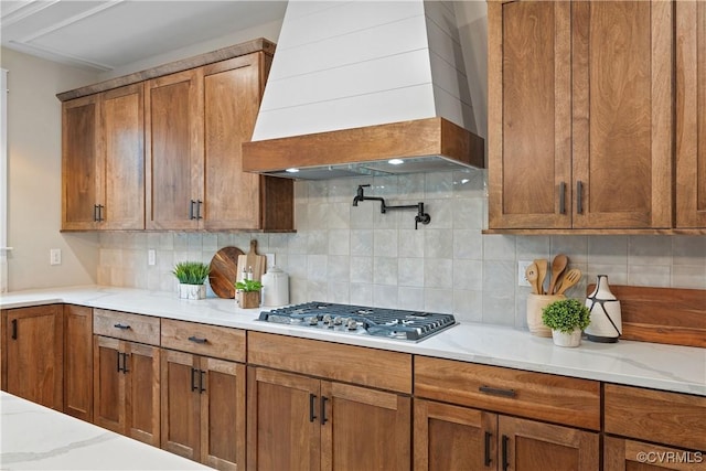 kitchen with custom range hood, light stone counters, decorative backsplash, stainless steel gas stovetop, and brown cabinetry