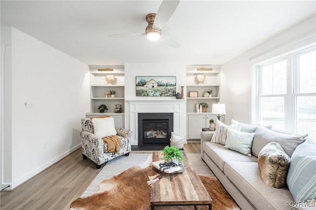 living room with built in shelves, baseboards, light wood-style flooring, a glass covered fireplace, and a ceiling fan