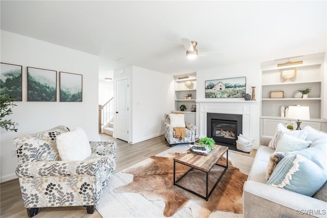 living area featuring baseboards, stairs, wood finished floors, a glass covered fireplace, and a ceiling fan