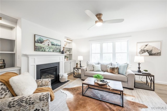 living area featuring a fireplace with flush hearth, built in shelves, a ceiling fan, and light wood finished floors