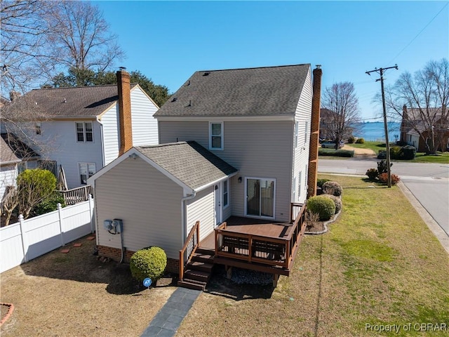 back of house featuring a lawn, a deck, fence, a shingled roof, and a chimney