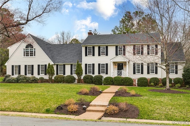 colonial inspired home featuring a chimney, a front yard, and a shingled roof