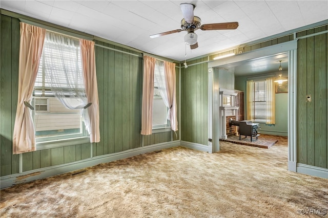 carpeted empty room with a fireplace, a ceiling fan, baseboards, and wood walls