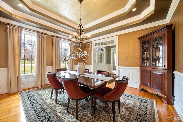 dining area featuring crown molding, wainscoting, light wood-style floors, an inviting chandelier, and a raised ceiling