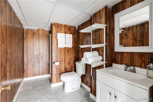 bathroom with a drop ceiling, wood walls, toilet, and vanity