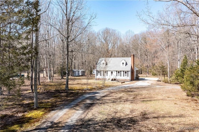 cape cod house featuring a porch, dirt driveway, an outdoor structure, a wooded view, and a chimney