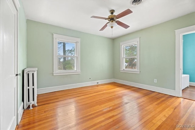 empty room with a ceiling fan, visible vents, baseboards, light wood-style flooring, and radiator heating unit