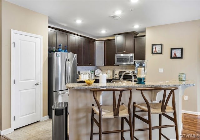 kitchen with a breakfast bar area, light stone counters, stainless steel appliances, decorative backsplash, and dark brown cabinets