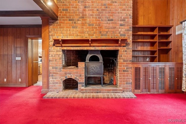 unfurnished living room featuring wooden walls, carpet flooring, beam ceiling, and a wood stove